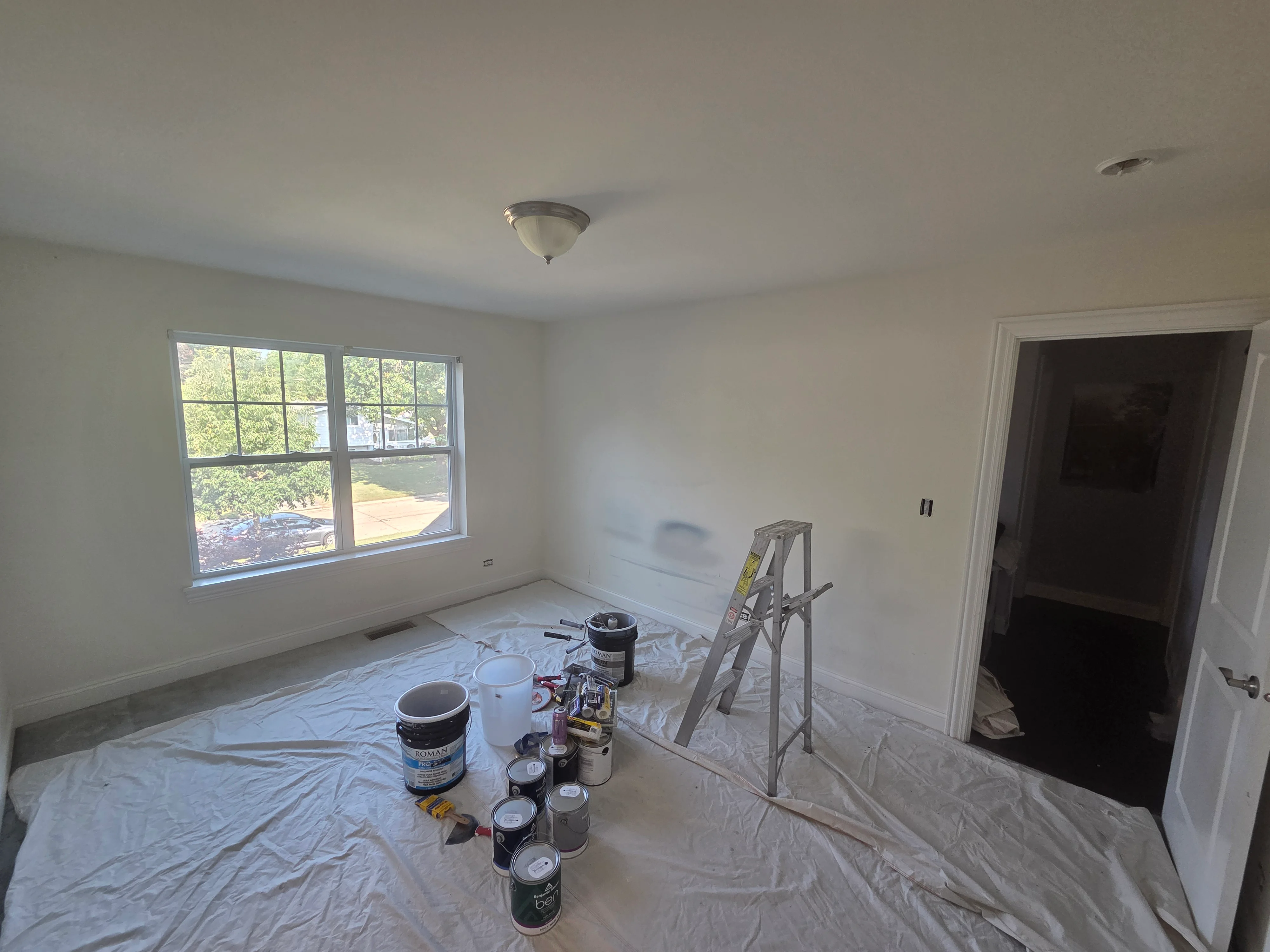 Interior room with primed drywall, drop cloths, paint cans, and ladder during painting preparation