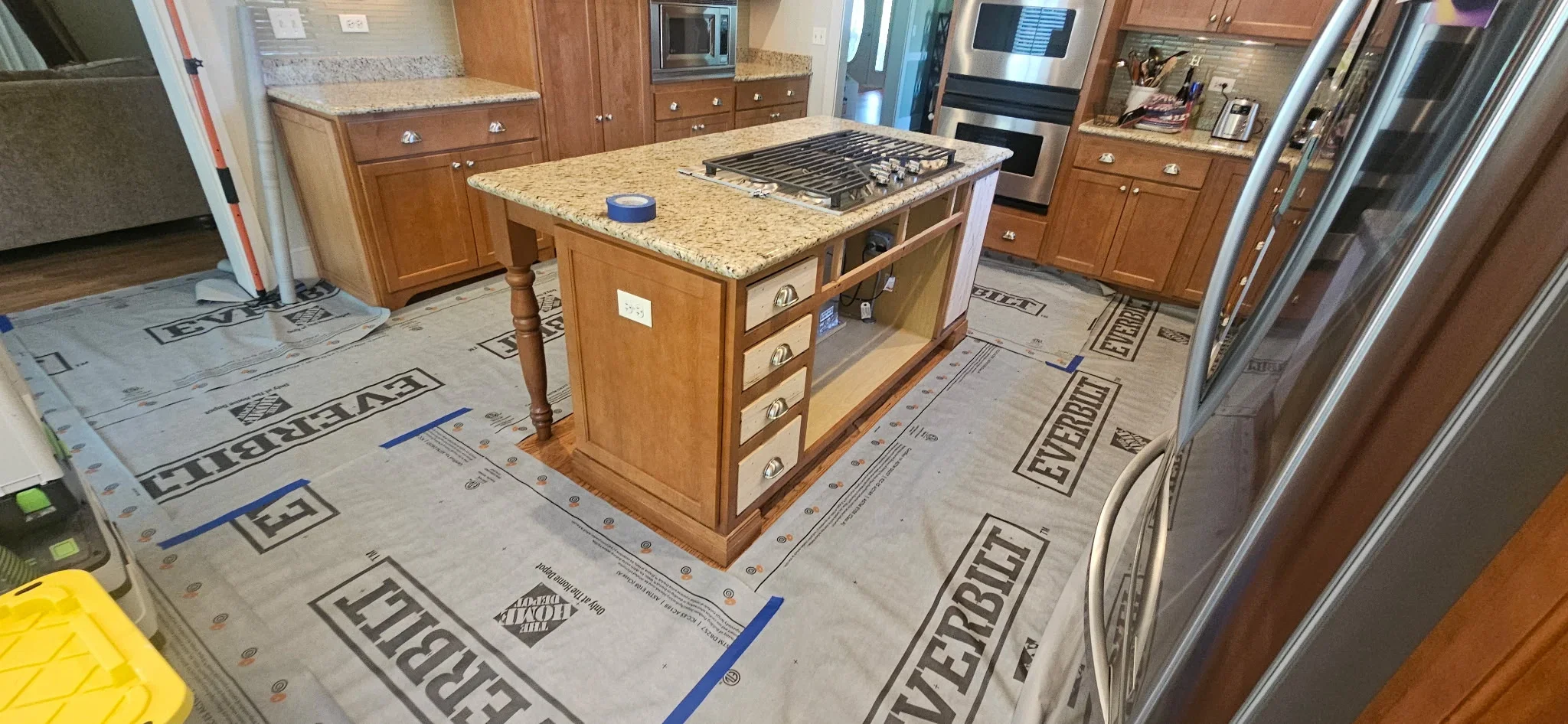 Kitchen with wood cabinets, granite counters, island with cooktop, and stainless steel appliances during renovation