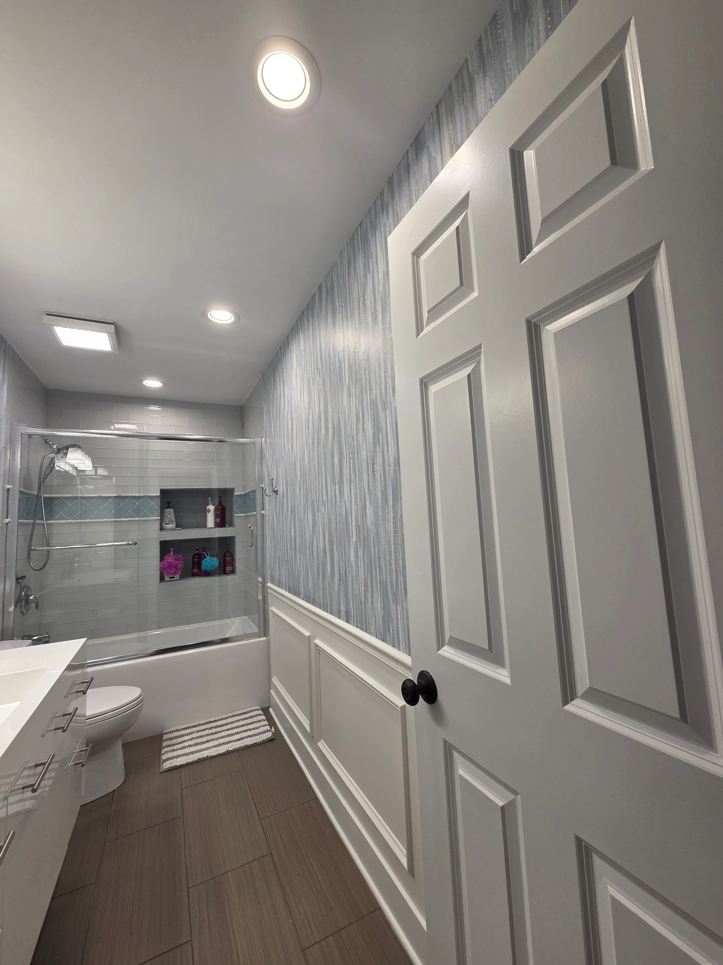 Bathroom with white painted walls, gray textured accent wallpaper, wainscoting, and recessed lighting