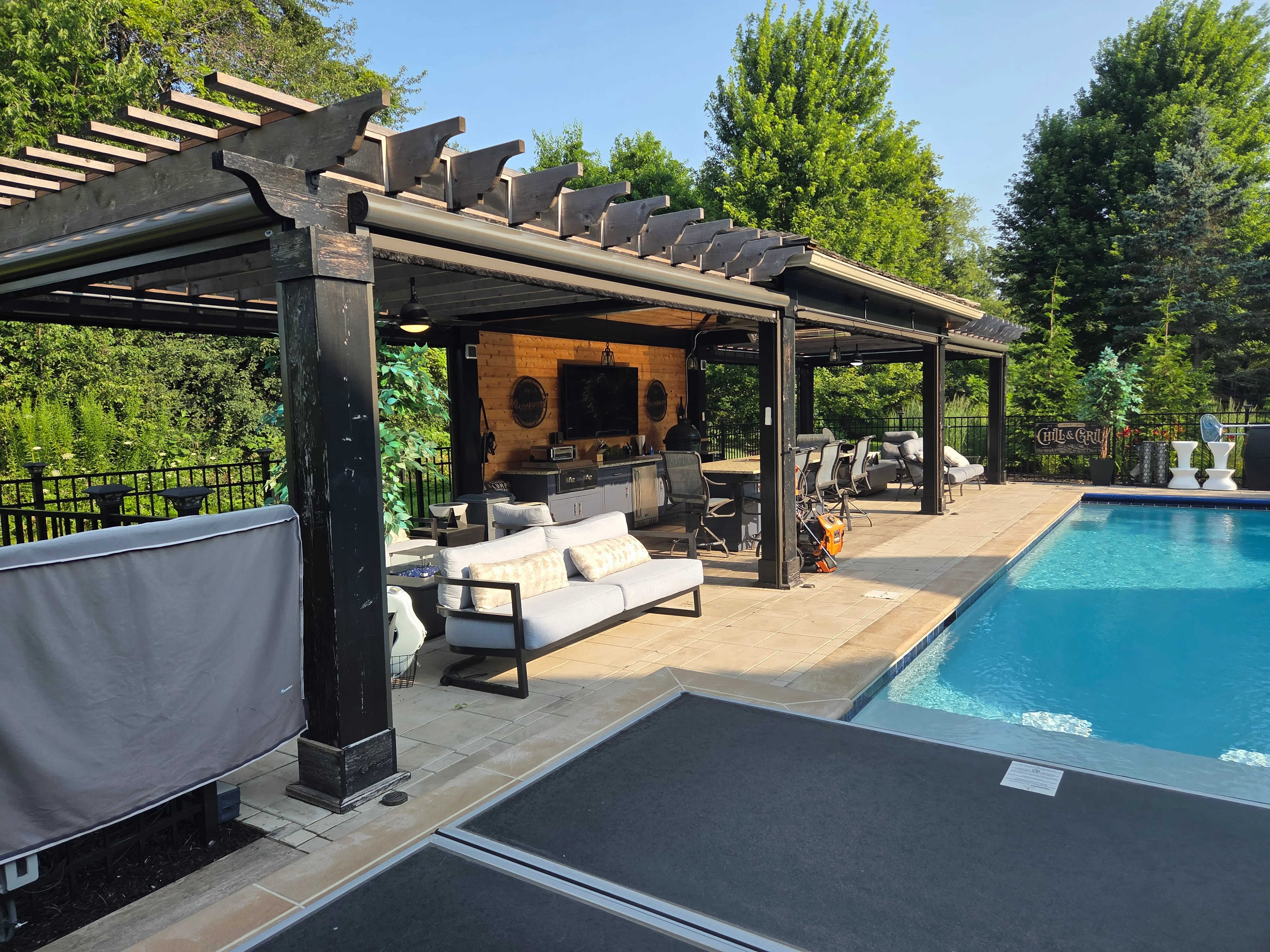 Black steel pergola with wood accent wall, TV installation, and outdoor seating by pool